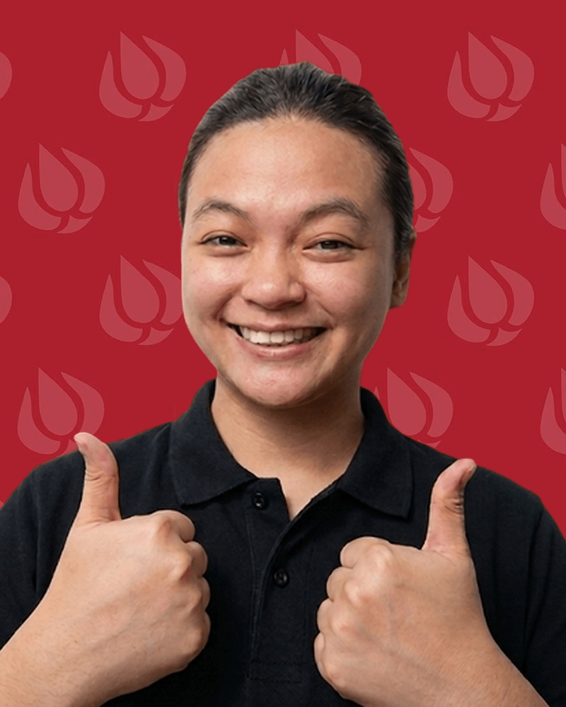 A person with long dark hair tied back, smiling and giving two thumbs up, wearing a black collared shirt—perfectly capturing the friendly spirit of an appliance repair company. The background is red with a repeating light pink abstract pattern.