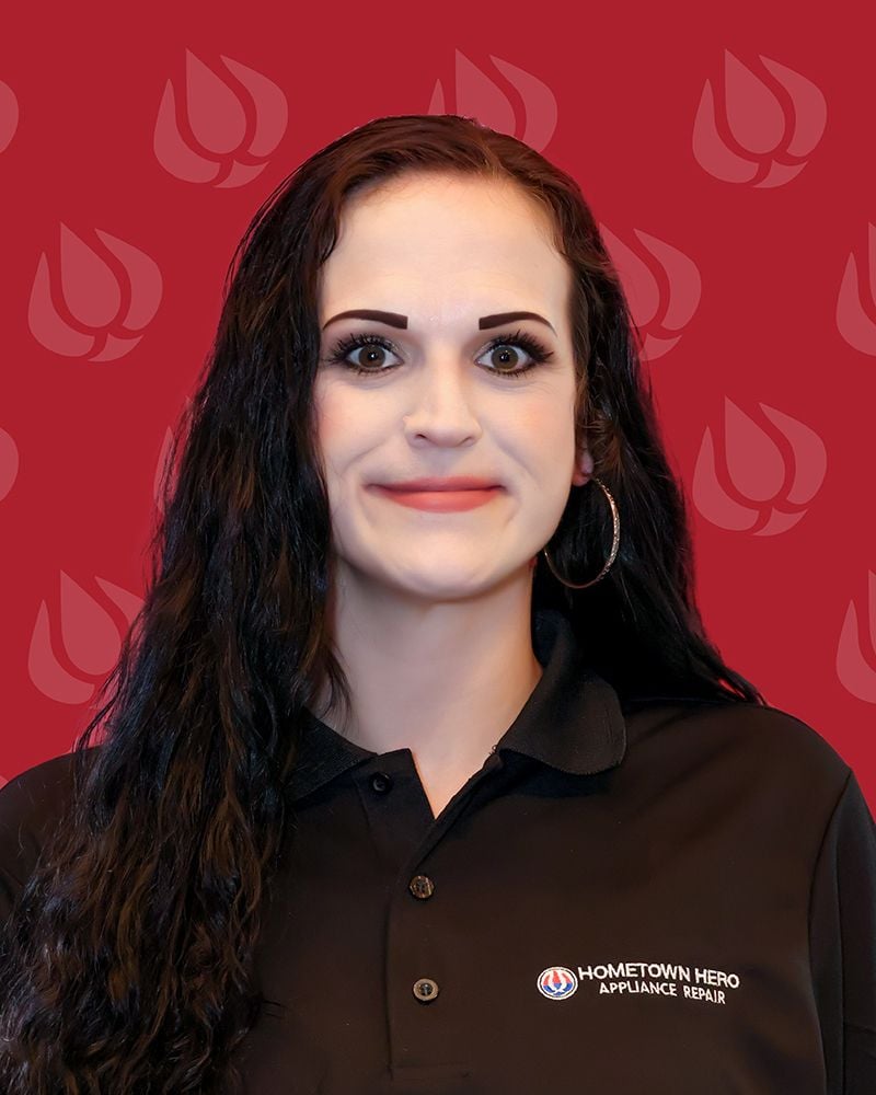 A woman with long dark hair and hoop earrings smiles in front of a red background with stylized flame logos, wearing a black "Hometown Hero Appliance Repair" polo shirt, representing a trusted local appliance repair company.