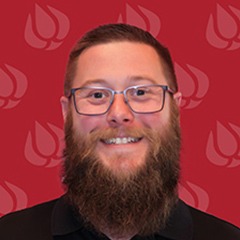 A smiling man with a full beard and glasses stands in front of a red background with a repeating flame logo pattern.