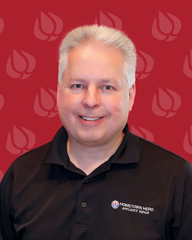A smiling man with short gray hair wearing a black Hometown Hero Appliance Repair polo shirt, representing an appliance repair company, stands in front of a red background with a repeating logo pattern.