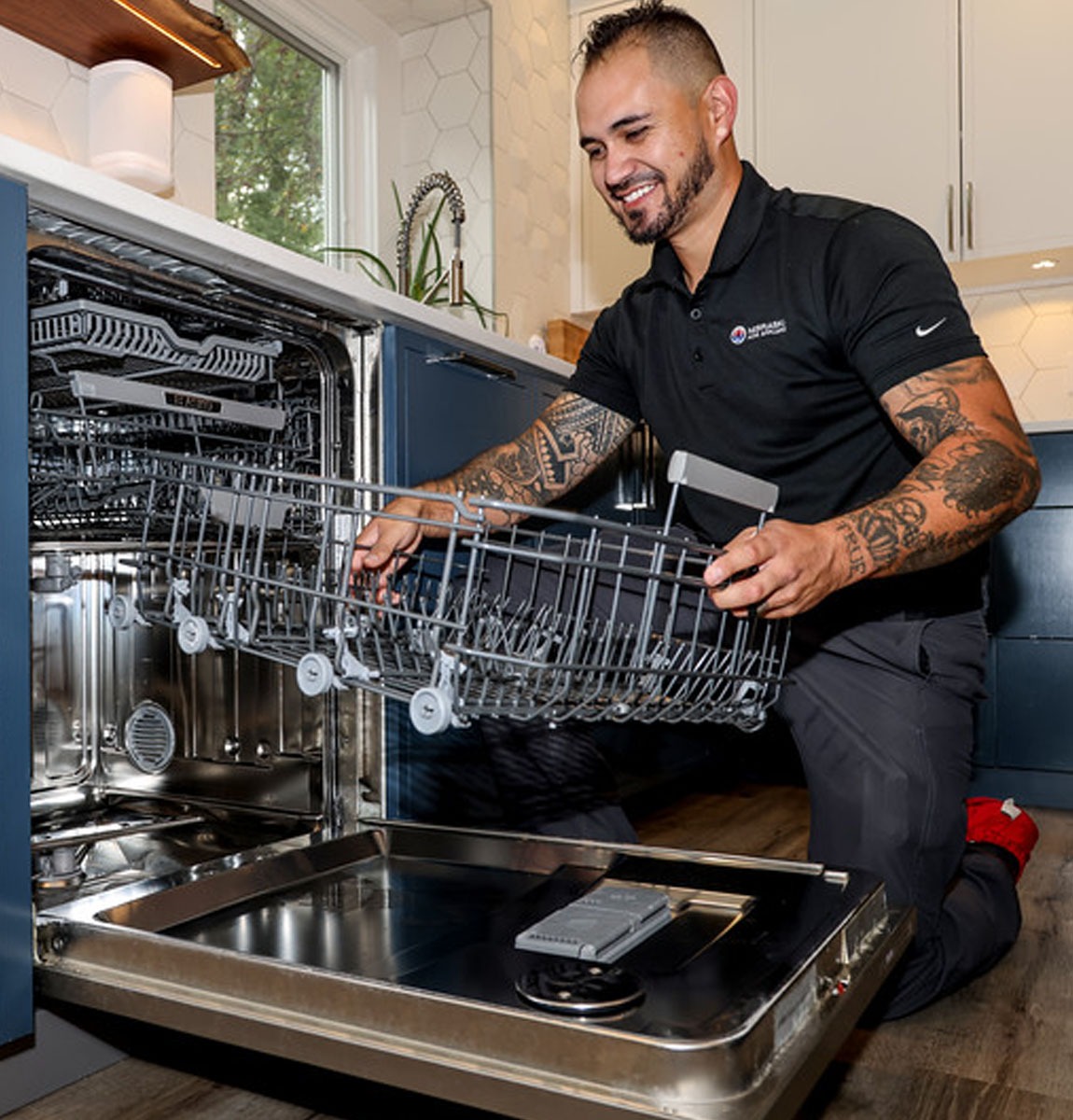 tech replacing dishwasher drawer A smiling man in a black shirt with tattoos inspects the lower rack of an open dishwasher in a modern kitchen. He is kneeling on the floor and appears to be performing maintenance or repairs.