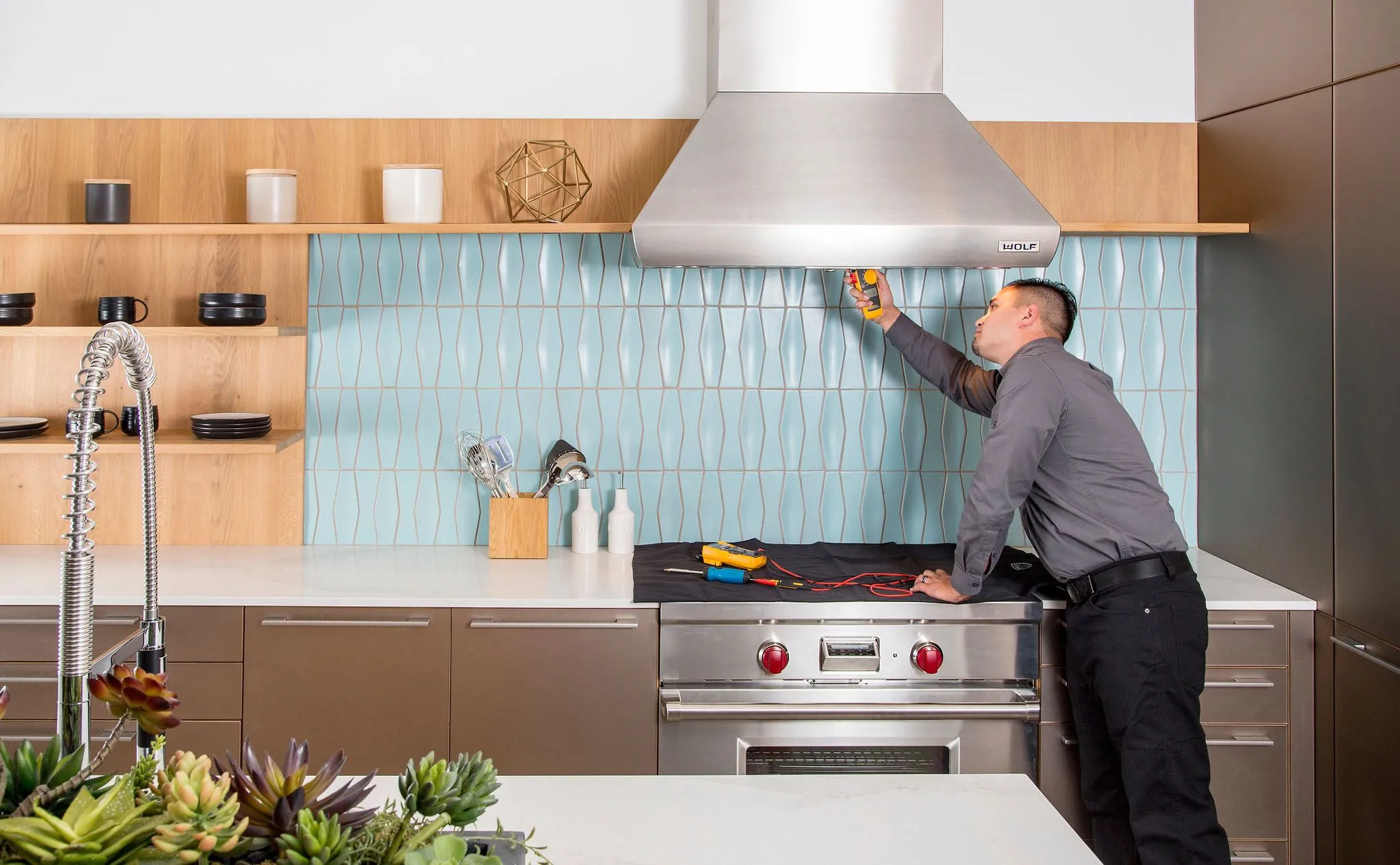 Tech testing range hood A man in a gray shirt inspects a stainless steel kitchen range hood with a handheld device in a modern kitchen with light blue tile backsplash and wooden shelves.
