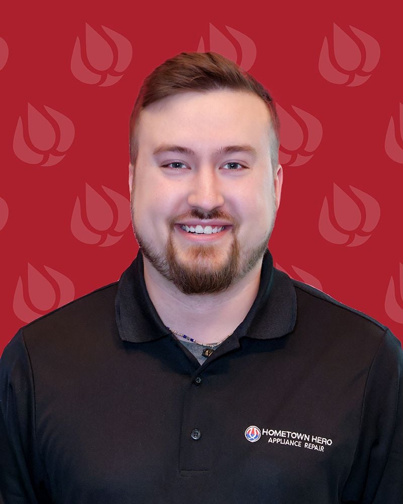 A smiling man with short brown hair and a trimmed beard, wearing a black polo shirt with the Hometown Hero Appliance Repair logo, stands in front of a red background featuring repeating logos for the appliance repair company.