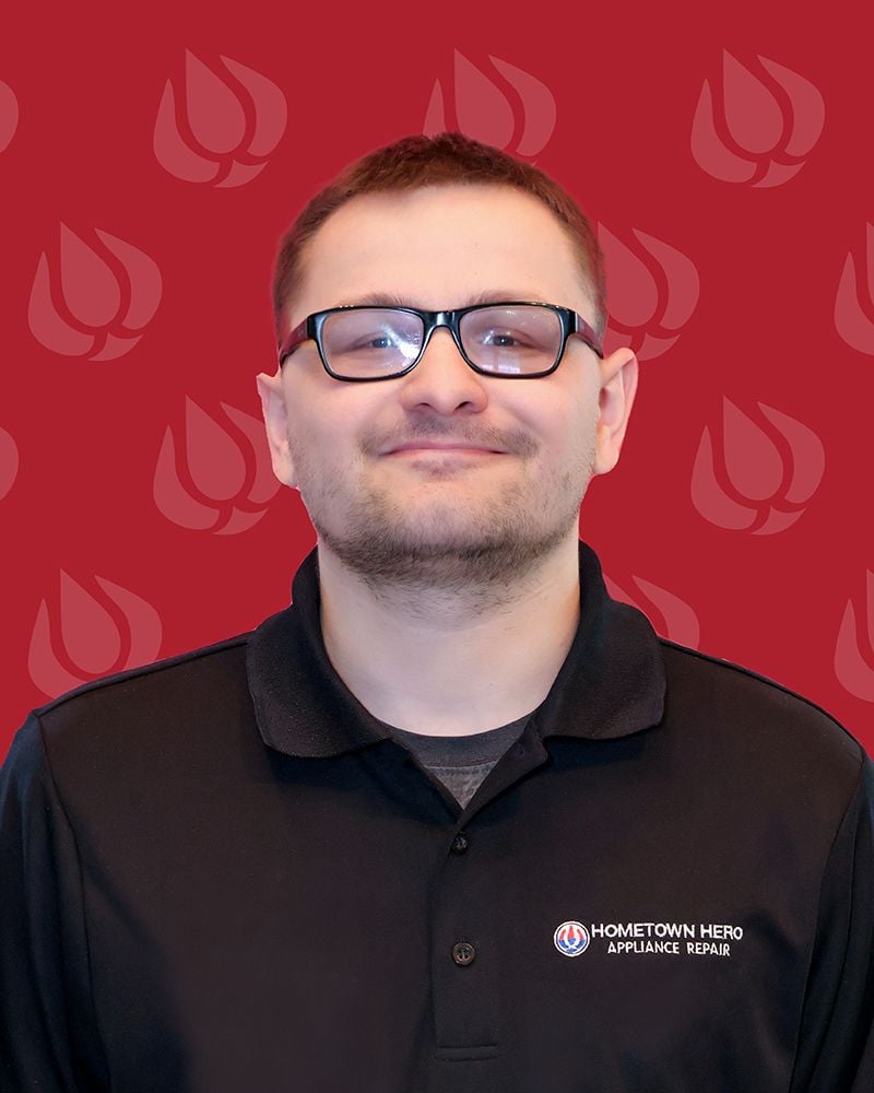 A person wearing glasses and a black Hometown Hero Appliance Repair shirt stands in front of a red background with a repeating appliance repair company logo pattern.