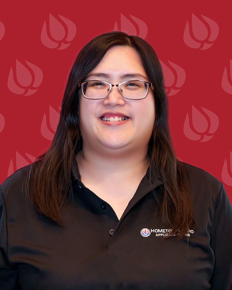 A woman with long brown hair and glasses smiles at the camera. She is wearing a black polo shirt with a Hometown Hero Appliance Repair logo, representing the trusted appliance repair company, standing in front of a red background with flame logos.