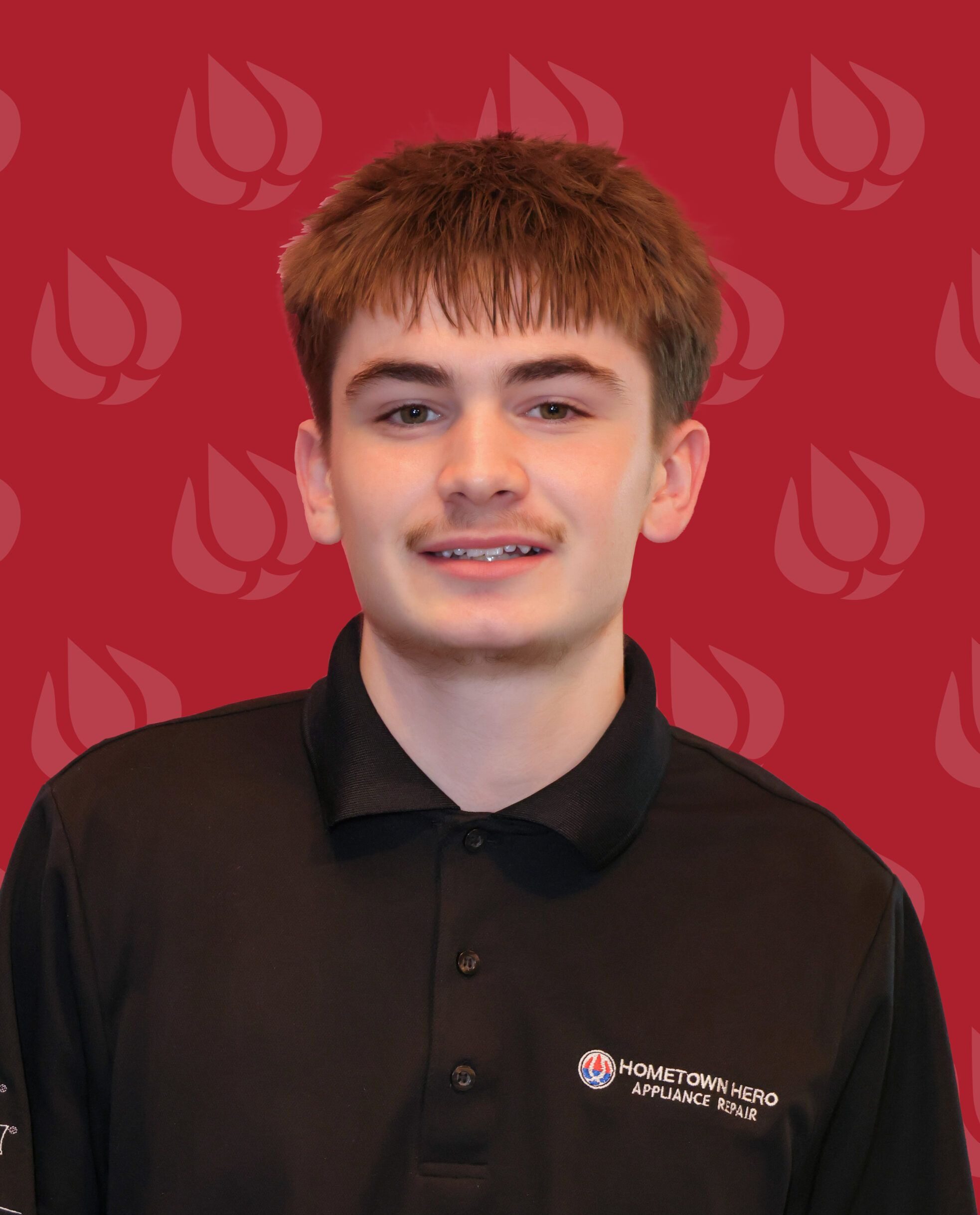 A young man with short brown hair wearing a black Hometown Hero Appliance Repair polo shirt stands in front of a red backdrop featuring his appliance repair company's repeating abstract flame logo.